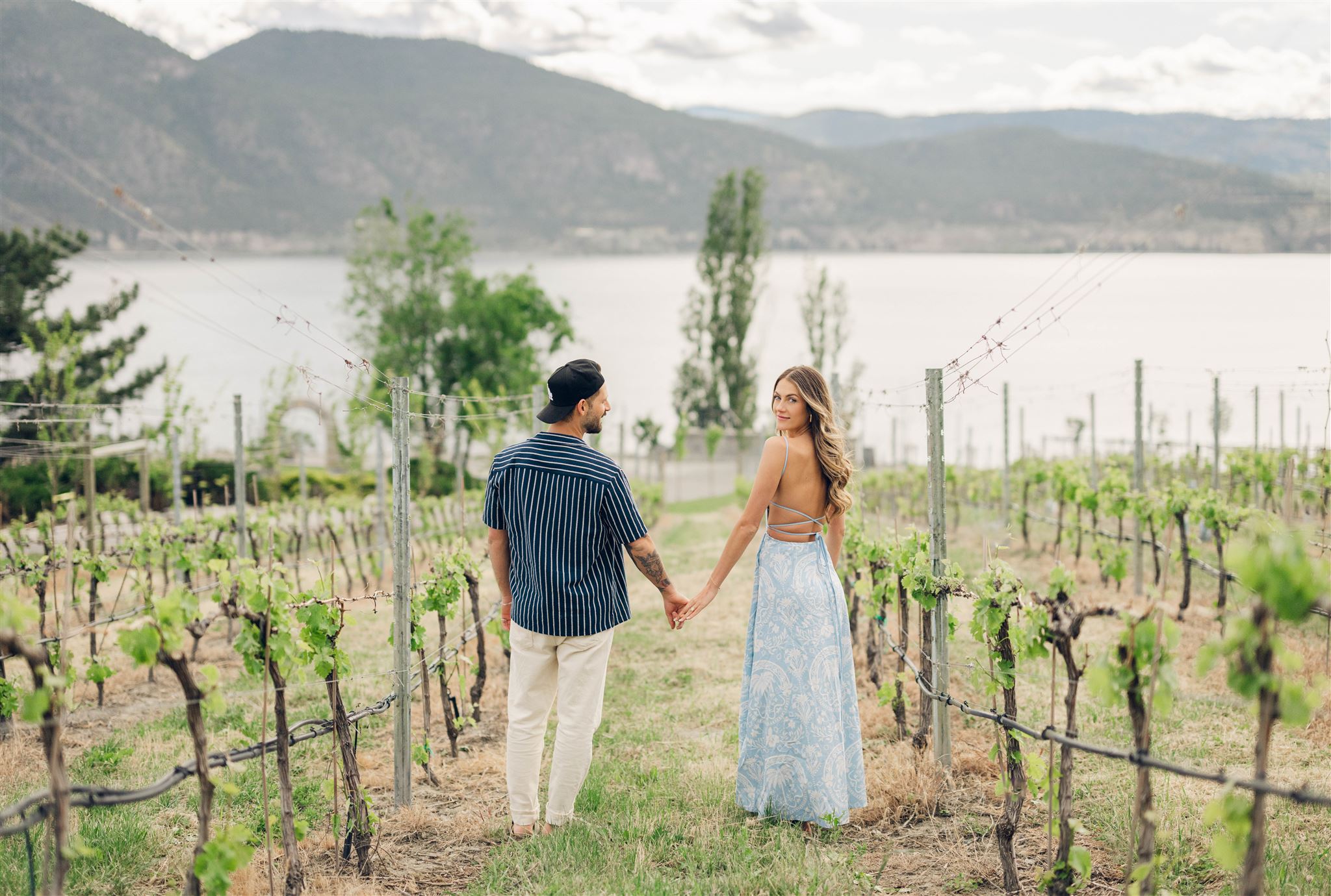 A couple standing in the vineyard at Bench 1775 Winery in Naramata during their engagement photo session with the views of mountains and lake in the background, after he proposed to her by surprise