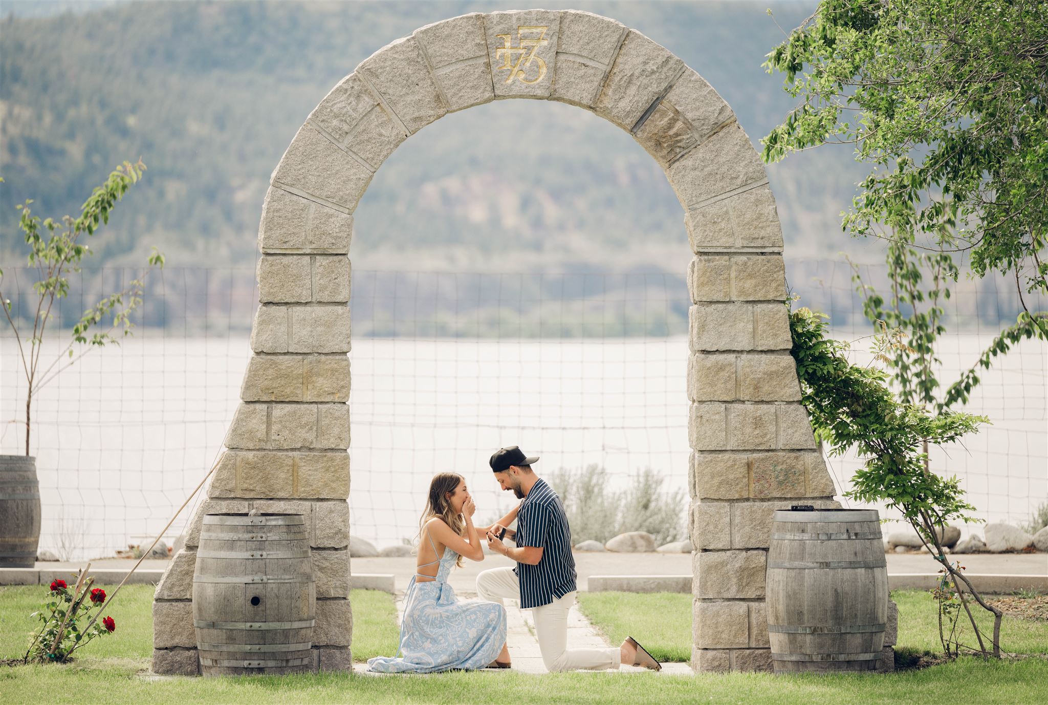 surprise marriage proposal, a man proposing to a woman in front of a stone arch with the lake and mountains in the background