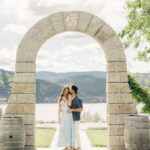 an engaged couple hugging under a stone arch with lake and mountains in the background after the marriage proposal